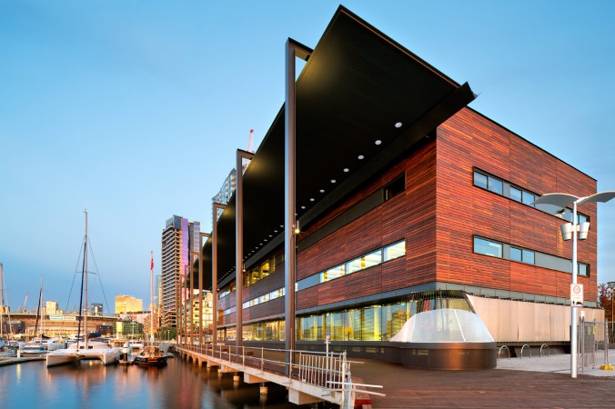 A modern building next to the bay, with boats in the background and a rosy sunset